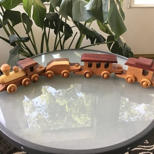 May include: A wooden toy train set with an engine, a boxcar, a flatcar, and a caboose. The train is made of different types of wood, including cedar and rosewood. The train is on a glass table top.