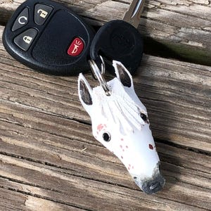 May include: A white leather horse head keychain with black and red painted details, including a black mane, black eyes, and red spots, attached to a black plastic car key fob with four buttons and a metal key blade on a silver metal ring.