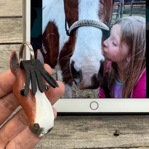 May include: A brown and white horse head keychain with a black leather mane and a silver metal ring. The keychain is held in front of a tablet screen displaying a photograph of a person kissing a brown and white horse on the nose.