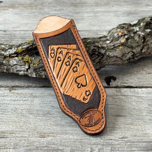 May include: A tan and dark brown leather sleeve featuring a stamped design of a royal flush of playing cards. The leather case has a rounded top opening and a circular stamp at the bottom that reads Cherokee Rose Custom Leather.