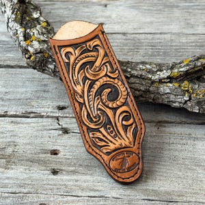 May include: A tan leather sheath featuring intricate hand-tooled floral scrollwork patterns and a dark brown stained background. The bottom of the sheath displays an oval stamp with the text Cherokee Rose Custom Leather and a feather icon.