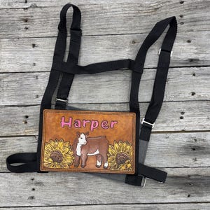 May include: A brown leather halter plate with the name "Harper" in pink lettering. The plate features a cow illustration with sunflowers. Black nylon straps and metal hardware complete the halter.