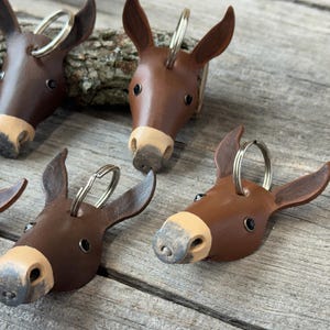 May include: A collection of brown leather donkey head keychains, each featuring a light tan muzzle, dark eyes, and a silver metal split ring attached to the top of the head. The keychains are shaped like donkey faces with pointed ears.