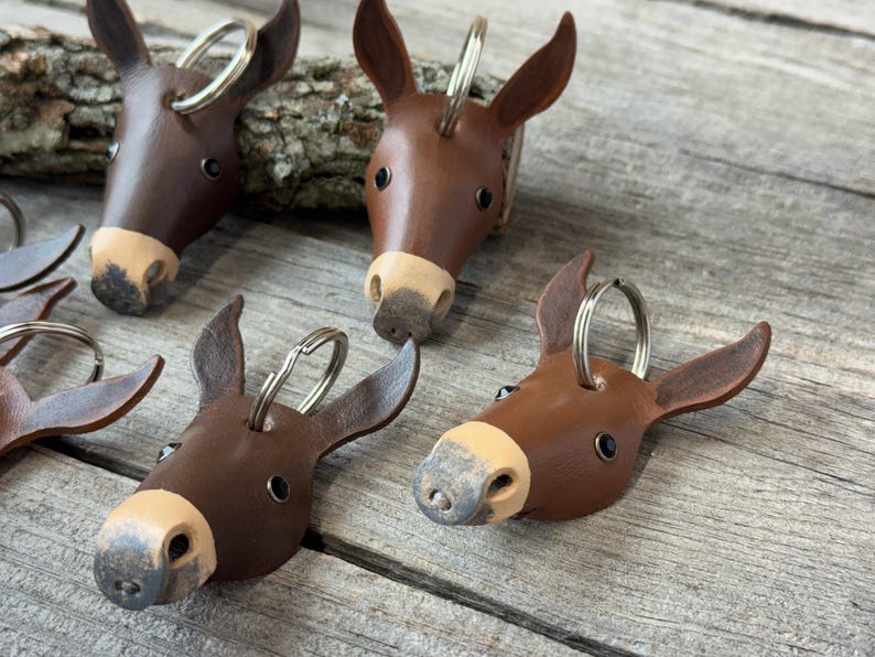 May include: A collection of brown leather donkey head keychains, each featuring a light tan muzzle, dark eyes, and a silver metal split ring attached to the top of the head. The keychains are shaped like donkey faces with pointed ears.