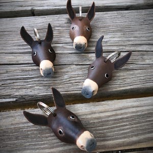 May include: Four handcrafted leather donkey keychains. Each keychain features a brown head with black ears and a light beige muzzle. The keychains are arranged on a weathered wooden surface.