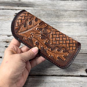May include: A brown leather case featuring an embossed pattern of oak leaves and acorns alongside a basketweave texture. The leather has a dark burnished edge and a curved top profile, showcasing detailed tooling across the entire surface.
