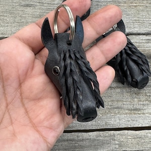 May include: A black leather keychain shaped like a horse head with a silver metal ring. The horse head features a dark, circular eye accent and a textured, braided mane made from thin strips of black leather attached to the side of the head.