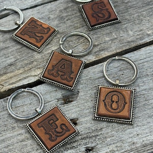 May include: Five square keychains featuring brown leather inserts with embossed letters N, S, A, O, and E. Each leather piece is set within a silver-toned metal frame with a beaded border and attached to a circular silver-toned metal split ring.