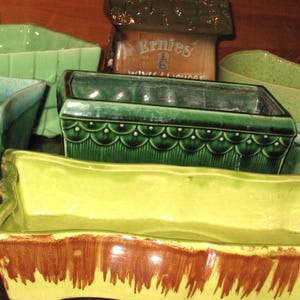 May include: A selection of vintage ceramic planters in shades of green and yellow. The planters have rectangular and curved forms, some with decorative patterns and brown trim. The image includes a small ceramic building with the text "Ernies Wine & Liquors".
