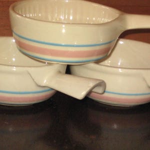 Pode incluir: Três tigelas de sopa de cerâmica branca com alças, empilhadas. Cada tigela tem uma faixa azul e rosa no meio. As tigelas estão dispostas de forma ligeiramente sobreposta, mostrando seu design.
