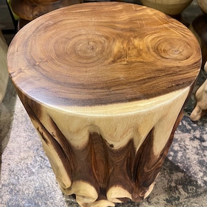 May include: A handcrafted wooden stool with a round top and a unique, carved base. The top is a rich brown with visible wood grain, while the base features a lighter, natural wood tone with a wavy, textured design. This decorative piece is a functional art.