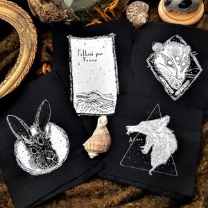 Puede incluir: Cuatro parches de tela negra con diseños bordados en blanco. Un parche presenta una ola con el texto "Follow your Fears". Otro parche presenta un conejo con tres ojos. El tercer parche presenta un lobo aullando a la luna. El cuarto parche presenta un mapache con tres ojos.
