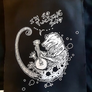 May include: Black patch with a white illustration of a badger playing a banjo. The badger is inside a crescent moon shape. Text above the badger reads "I'M SO FUCKING SAD".