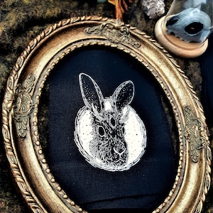 Peut inclure: Un patch en tissu noir avec une illustration blanche d'un lapin à trois yeux et des cornes. Le lapin est entouré d'un cercle d'étoiles. Le patch est encadré dans un cadre ovale doré.