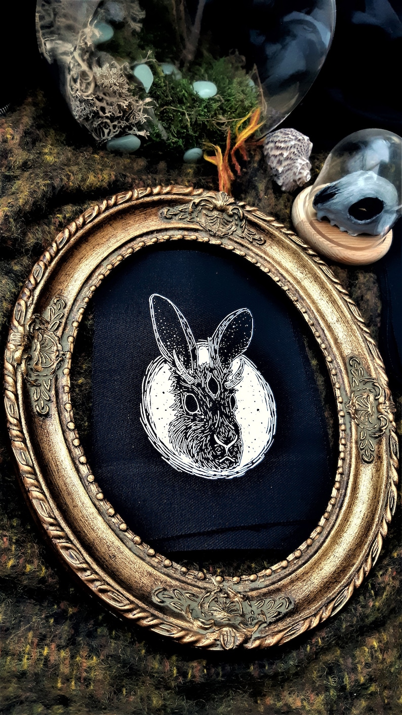 May include: A black fabric patch with a white illustration of a rabbit with three eyes and horns. The rabbit is surrounded by a circle of stars. The patch is framed in a gold oval frame.