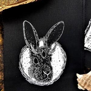 May include: Black and white embroidered patch featuring a stylized rabbit with three eyes and horns, set against a white circle on a black fabric background.