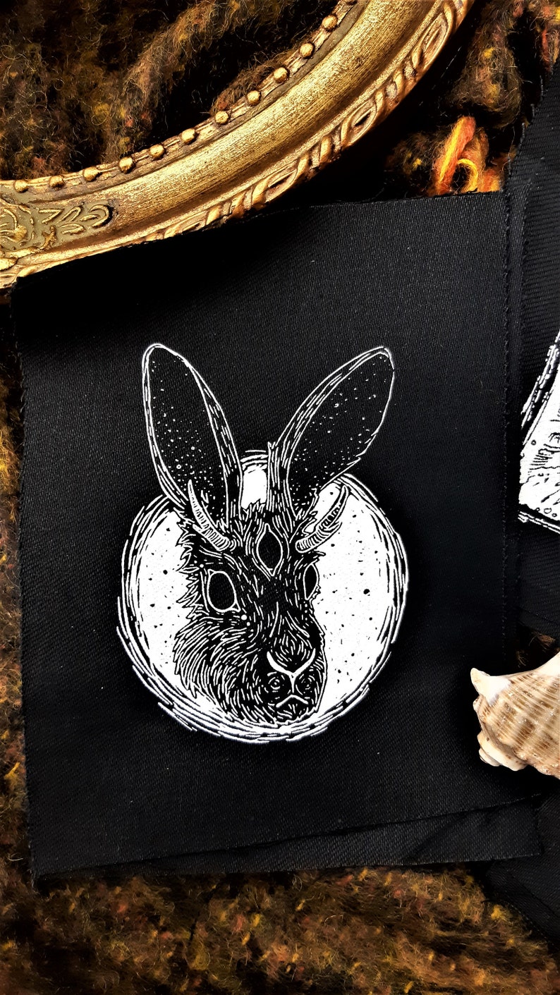 May include: Black and white embroidered patch featuring a stylized rabbit with three eyes and horns, set against a white circle on a black fabric background.