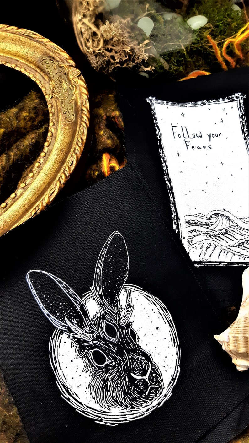 May include: Black and white embroidered patch featuring a rabbit with three eyes and antlers, surrounded by a circle. The patch is on a black fabric background. The text "Follow your Fears" is written on a separate white patch with a black border.
