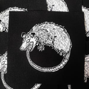 Könnte beinhalten: Schwarzer quadratischer Aufnäher mit einer weißen Illustration eines Opossums, das sich in den Schwanz beißt. Das Opossum ist mit detailliertem Fell und scharfen Zähnen dargestellt. Mehrere Aufnäher sind gestapelt und zeigen das gleiche Design.