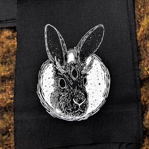 May include: Black and white illustration of a rabbit with four eyes, printed on a black fabric patch.