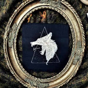 May include: A black fabric patch with a white embroidered image of a wolf howling inside a triangle. The wolf is stylized and has a starry background. The text "ARAAA" is embroidered below the wolf.