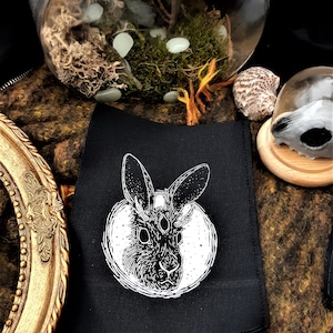 May include: Black fabric patch with a white illustration of a rabbit with three eyes and antlers. The rabbit is surrounded by a circle with a starry background.