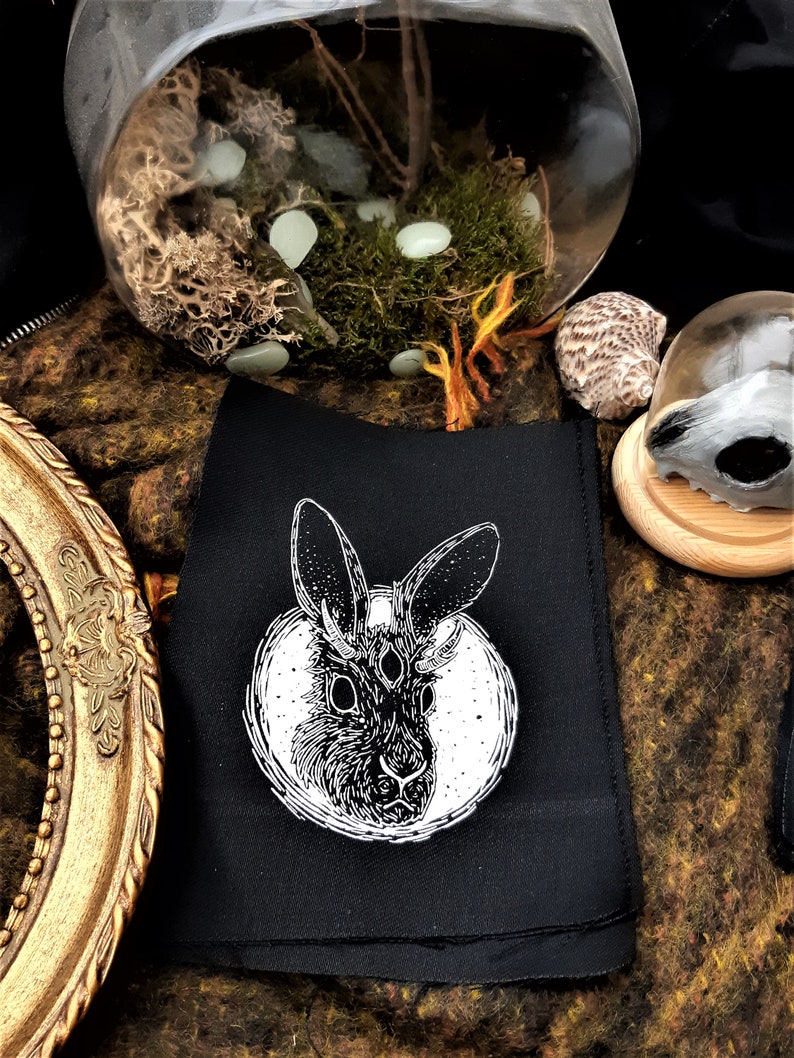 May include: Black fabric patch with a white illustration of a rabbit with three eyes and antlers. The rabbit is surrounded by a circle with a starry background.