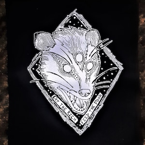 May include: Black patch featuring a stylized opossum illustration with three eyes, set within a diamond shape. The text "EAT THE RICH" and "FEED THE POOR" are printed on the border. The opossum is in shades of gray and white.
