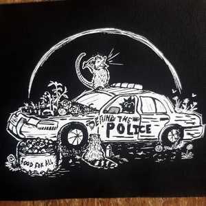 May include: Black and white graphic patch featuring a police car with the words "Defund the Police." A rat sits on top of the car, and a raccoon is in front. The car's hood is a garden with the words "Food for All" on a bucket.