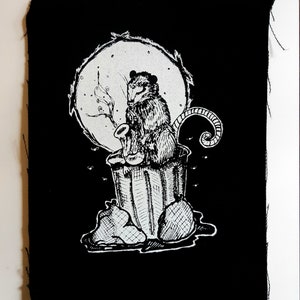Pode incluir: Ilustração em preto e branco de um gambá tocando saxofone em um lixo. O gambá está sentado na borda do lixo, com o rabo enrolado ao redor do lado. O lixo está cheio de lixo e há moscas zumbindo ao redor. O gambá está usando um chapéu e óculos de sol, e tem uma expressão travessa.