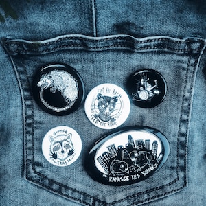 May include: A collection of five circular and oval pin-back buttons featuring black and white illustrations. Designs include an opossum, a raccoon labelled TRASH, a drummer, a raccoon with the text EAT THE RICH FEED THE POOR, and pigeons with the text RAMASSE TES BITCHES.