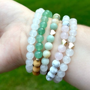 May include: A stack of five bracelets with different colored beads. The bracelets are made of white, green, and pink beads. The beads are round and have a smooth finish. There are small gold heart charms on the bracelets.