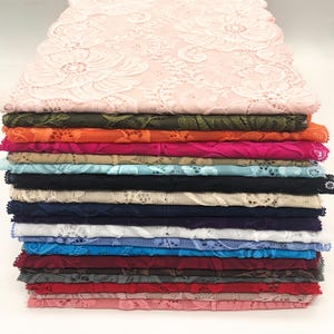 May include: A pile of colourful lace fabric in various shades. The top layer is a pale pink with a floral pattern. Beneath are layers of green, orange, pink, beige, blue, black, navy, and other colours. The lace has a delicate, open weave.