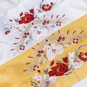 Puede incluir: Tela blanca con flores rojas y conejos blancos bordados. La tela está decorada con un patrón repetitivo de flores y conejos.