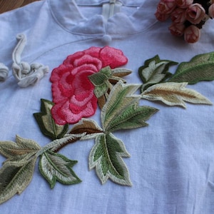 Könnte beinhalten: Ein weißes Leinenhemd mit einer gestickten rosa Blume und grünen Blättern. Die Blume ist eine Pfingstrose und die Blätter sind mit goldenen Nähten verziert.