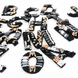 May include: Black, white, and gold beaded letters scattered on a white surface. The letters are embellished with beads and sequins, creating a sparkling effect. The letters are in various fonts and sizes.