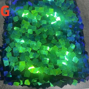 May include: Close-up of a fabric covered in square sequins. The sequins transition from green to blue, creating an ombre effect. The letter "G" is visible in the upper left corner. The sequins are attached to a black mesh backing.