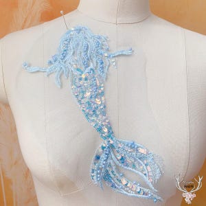 May include: A light blue mermaid applique with detailed bead and sequin embellishments. The mermaid features flowing hair, outstretched arms, and a detailed tail. Ideal for crafting and embellishment projects.