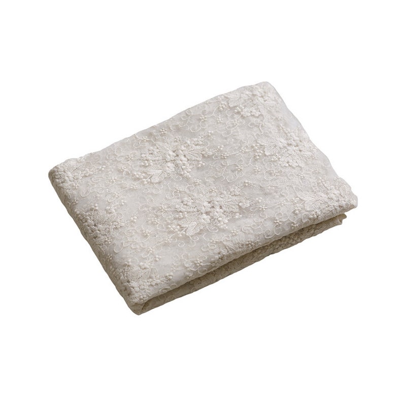 May include: A white lace fabric with a delicate floral pattern. The fabric is folded in half and has a soft, flowing texture.
