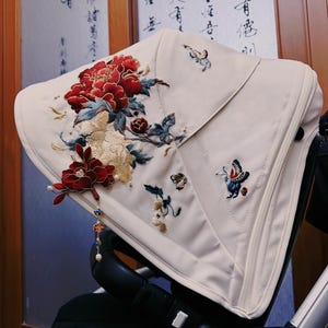 Op de afbeelding: Crèmekleurige kinderwagenkap met ingewikkelde bloemen- en vlinderborduursels in rood, crème, blauw en goud. Het ontwerp toont een grote pioenroos en delicate vlinders, met een decoratieve bedel aan de zijkant.