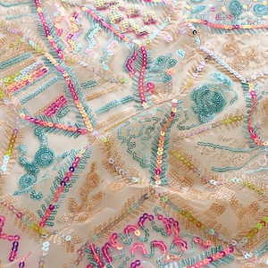 May include: A close-up of a sheer fabric with a colorful geometric pattern made of sequins. The fabric is a light beige color and the sequins are in shades of pink, blue, green, and gold.