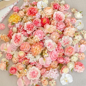 Può includere: Una collezione di fiori artificiali in varie tonalità di rosa, pesca, giallo e bianco. L'assortimento comprende rose, orchidee e altre varietà floreali, alcune con accenti di perle. La disposizione è sparsa, creando un'esposizione visivamente accattivante.