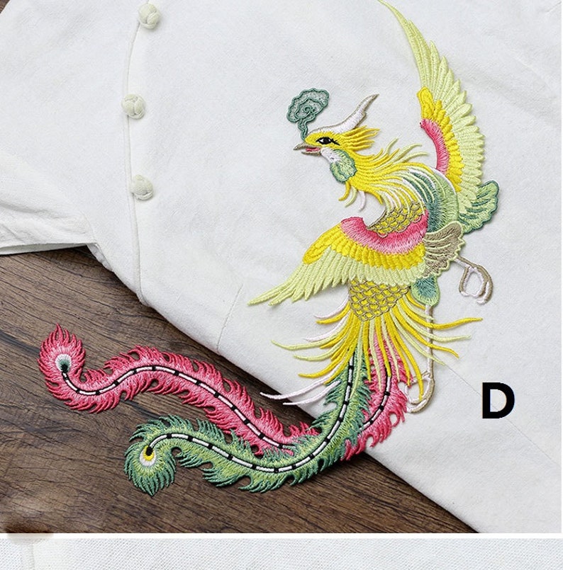Op de afbeelding: Twee geborduurde feniksen met gele, groene, roze en witte veren. De feniksen bevinden zich op een witte stoffen achtergrond. De letter "D" bevindt zich in de rechterbenedenhoek van de afbeelding.