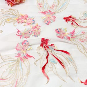 May include: Embroidered fabric with koi carp designs in red, pink, and gold. The fabric has a white background, and the fish are arranged in a flowing pattern. The fish have delicate details, with flowing tails and fins.
