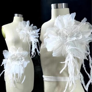 May include: Two white floral appliques on dress forms. The appliques feature layered fabric petals, a central flower, and long, flowing feathers. The design is delicate and elegant, suitable for bridal or formal wear.