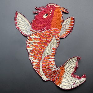 以下が含まれることがあります： 赤と白の刺繍されたコイの魚のワッペン。魚は大きく、口を開けた詳細な頭と大きな白いヒレを持っています。