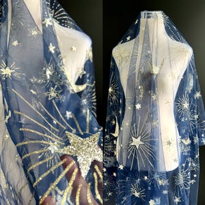 May include: Navy blue tulle fabric with a celestial design. The fabric features gold and silver glitter stars, moons, and bursts of light. The fabric is draped over a mannequin, showcasing the design and texture.