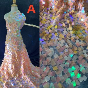 May include: A dress form draped with iridescent fabric. The fabric is covered in small, square sequins in shades of peach, yellow, and white. The sequins catch the light, creating a shimmering effect. The letter "A" is visible in the upper left corner.
