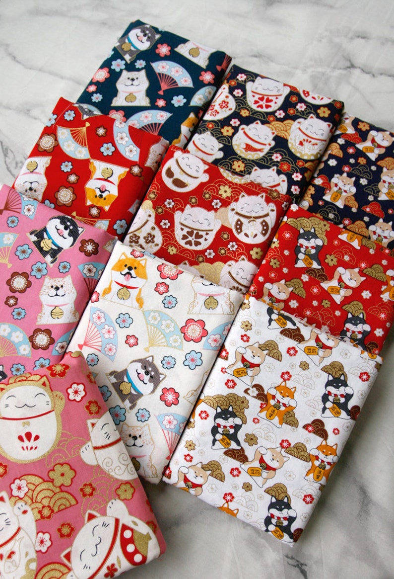 Puede incluir: Varias piezas de tela dobladas con estampados de inspiraci&oacute;n japonesa. Las telas presentan dise&ntilde;os repetidos de gatos de la suerte, perros Shiba Inu, abanicos y motivos florales en colores como rojo, rosa, azul y blanco.