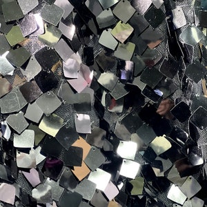May include: Close-up of a fabric covered in small, square, silver sequins. The sequins are attached to a black mesh backing. The sequins reflect light, creating a shimmering effect. The overall texture is dense and visually striking.